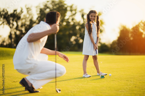 The father squats with a golf club in his hand and looks at his daughter, who looks at him and prepares to hit the ball