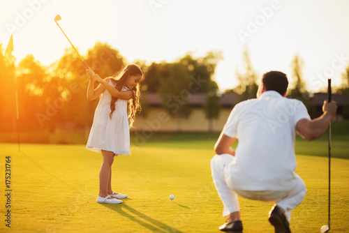 A man is squatting on a golf club and looking at a girl who is swinging a club to strike the ball