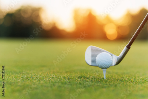 The golf club behind the golf ball on the stand. Against the background of grass and sunset