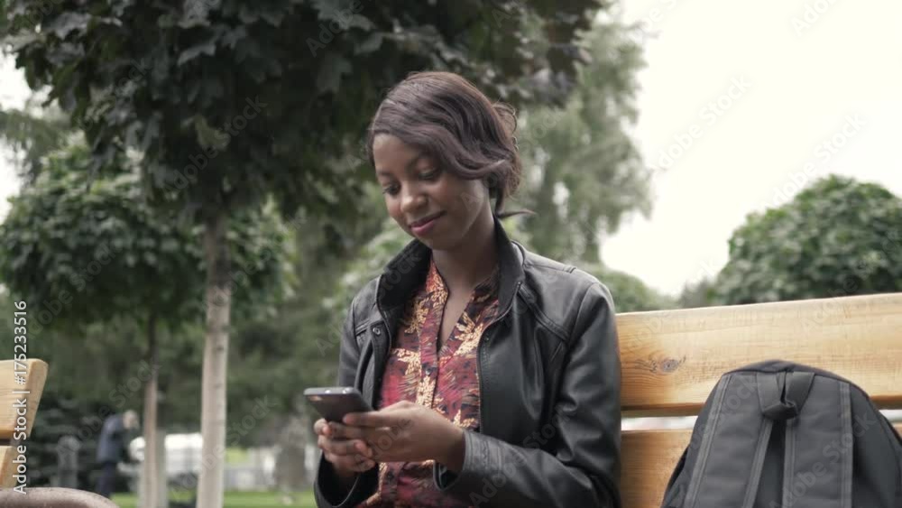 Portrait of beautiful young african woman smiling while sitting outside on a bench holding mobile phone after university, student girl Portrait of beautiful young african woman smiling while sitting outside on a bench holding mobile phone after university, student girl