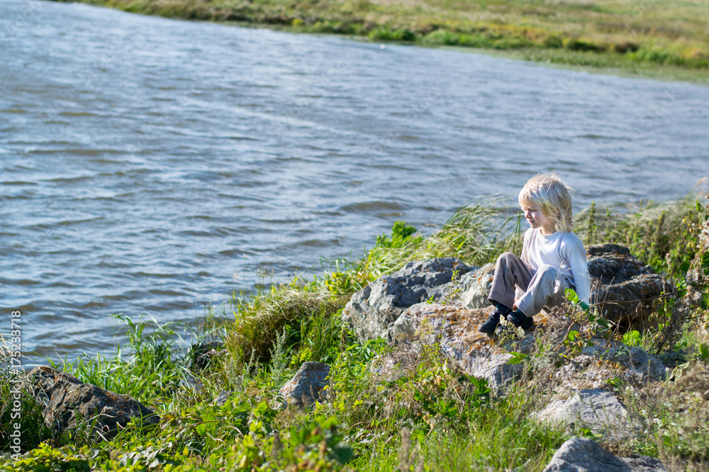 Blond thoughtful boy sitting at a river bank on a rocks - Rusiian Urals wind sunny day