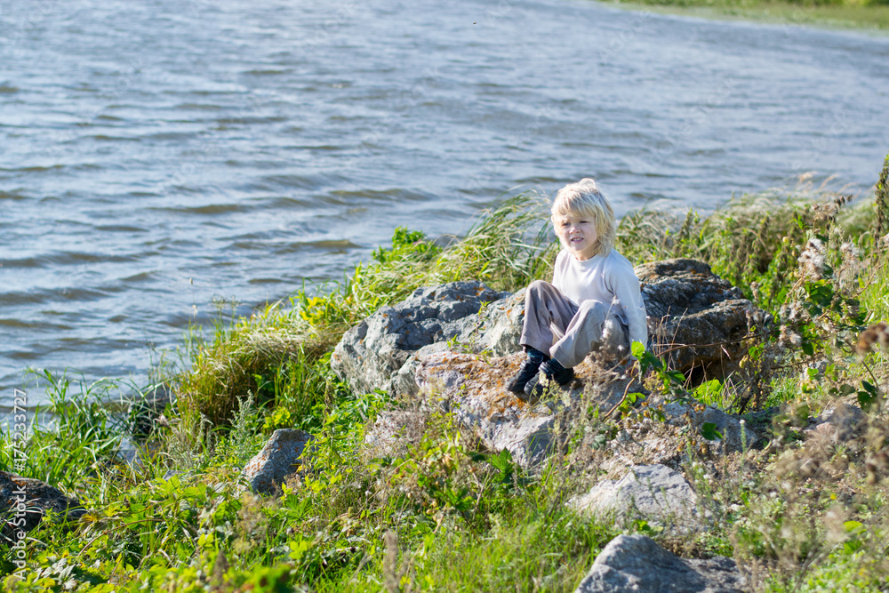 Blond thoughtful boy sitting at a river bank on a rocks - Rusiian Urals wind sunny day