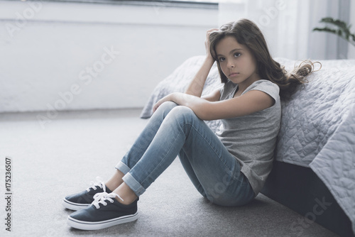 Fotografía A small dark-haired girl sits on the floor leaning on the bed and propping her h