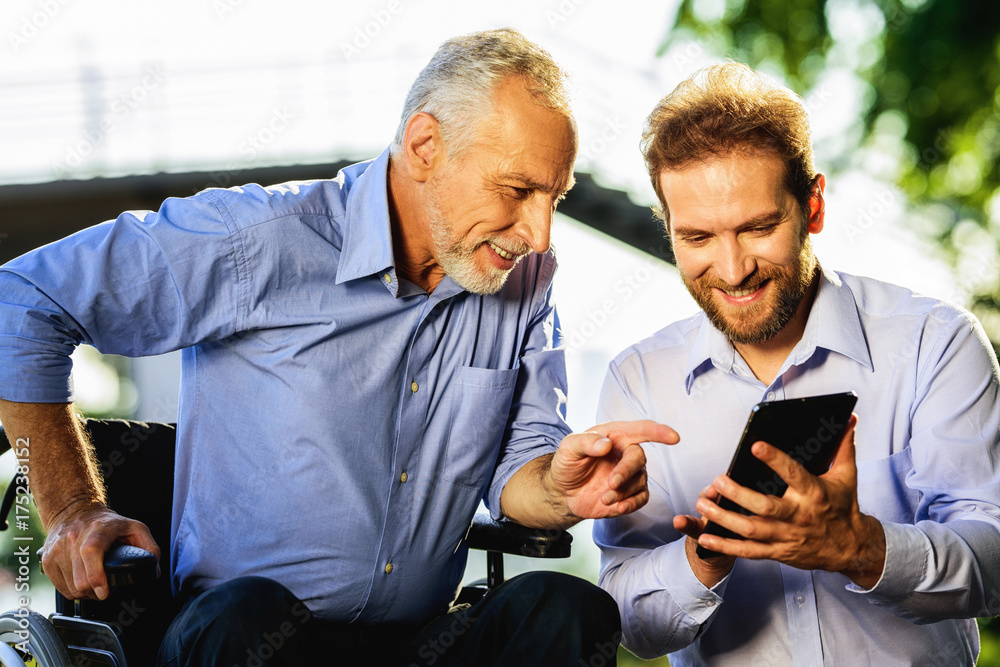 Old man in a wheelchair and a man looking at something in a tablet walking in the park. Old man shows something to a man Old man in a wheelchair and a man looking at something in a tablet walking in the park. Old man shows something to a man