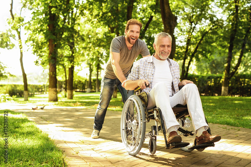 A pensioner on a wheelchair and his adult son are walking around the park. They are happy and have fun