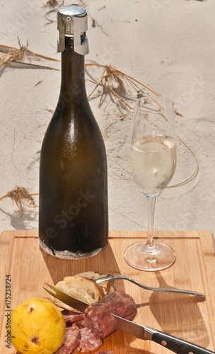 Fototapeta Naklejka Na Ścianę i Meble -  Chilled glass and bottle of vintage champagne, french bread, slices of salami and a pear on a bamboo serving plate on the sand of a tropical beach celebrating the autumn season