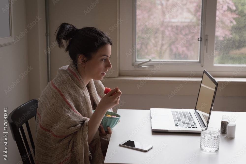 Woman having fruit while looking into laptop at table Woman having fruit while looking into laptop at table