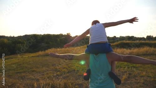 Little boy sitting on the shoulders of his father and playing airplane. Daddy carrying son and raised hands as plane. Family spending time together at nature. Slow motion Rear vack view photo