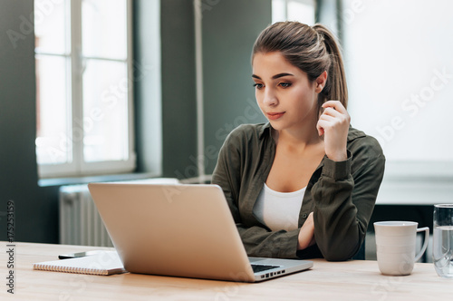 Young blond female student working on a laptop concentrating on reading the screen.