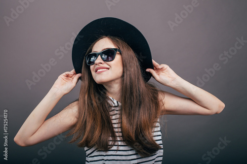 Young beautiful fashionable woman with trendy makeup in black hat and glasses on the grey background . Model looking at camera, wearing stylish eyeglasses. Female fashion, beauty concept. 