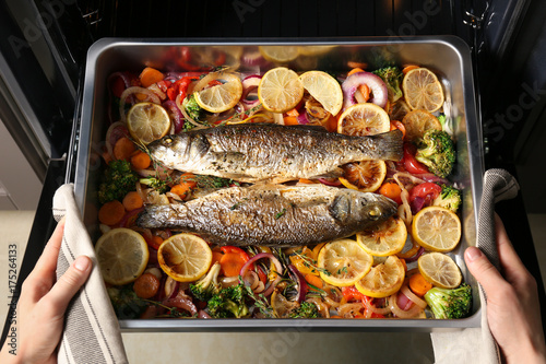 Woman taking baking tray with fish and vegetables out of oven