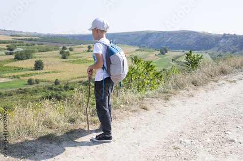 Small foot tourist. The boy dressed of black trousers , white t-shirt and cap with grey and blue backpack. Concept of travel in mountain. Child relying on a stick, staring into the distance.