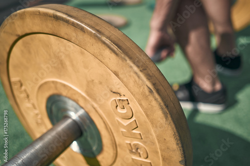 Close-up. Athletic man holding a bar with weights. Outdoors. Green colors. Sports ground.