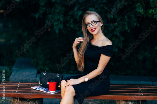 Young business lady in black dress and glasses with coffee and a bag sitting on bench in the park