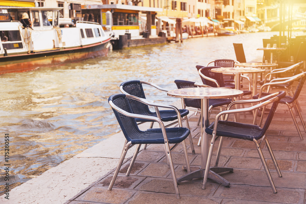 Fototapeta premium Restaurant tables and chairs with vaporetto ships in the background in Venice, Italy. European travel, outdoor dining and cuisine.