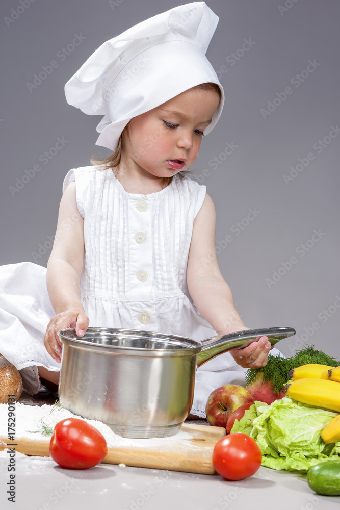 Coocking Ideas and Concepts. Portrait of Caucasian Little Girl In Cook Uniform Working With Saucepan In Studio with Vegetables At Background. Posing Against Gray. Coocking Ideas and Concepts. Portrait of Caucasian Little Girl In Cook Uniform Working With Saucepan In Studio with Vegetables At Background. Posing Against Gray.