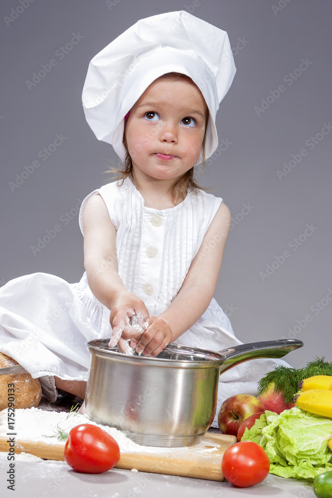 Coocking Ideas and Concepts. Portrait of Surprised Caucasian Little Girl In Cook Uniform With Saucepan In Studio with Vegetables At Background. Posing with Dreaming Facial Expression. Against Gray. Coocking Ideas and Concepts. Portrait of Surprised Caucasian Little Girl In Cook Uniform With Saucepan In Studio with Vegetables At Background. Posing with Dreaming Facial Expression. Against Gray.