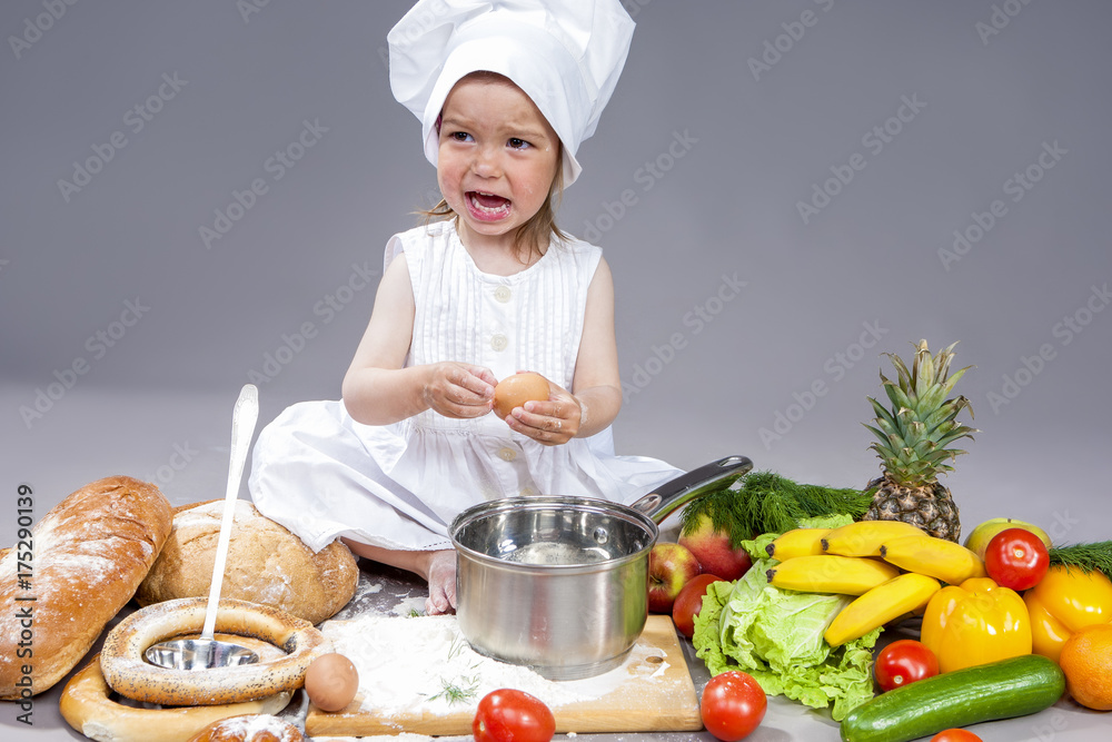 Surprised Caucasian Little Girl In Cook Uniform With Fresh Eggs and Saucepan In Studio with Vegetables At Background. Posing with Angry Expression. Against Gray. Surprised Caucasian Little Girl In Cook Uniform With Fresh Eggs and Saucepan In Studio with Vegetables At Background. Posing with Angry Expression. Against Gray.