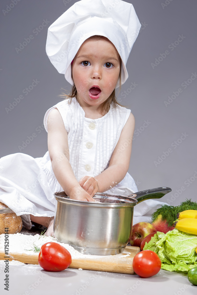 Coocking Ideas and Concepts. Portrait of Surprised Caucasian Little Girl In Cook Uniform With Saucepan In Studio with Vegetables At Background. Posing with Mouth Open. Against Gray. Coocking Ideas and Concepts. Portrait of Surprised Caucasian Little Girl In Cook Uniform With Saucepan In Studio with Vegetables At Background. Posing with Mouth Open. Against Gray.