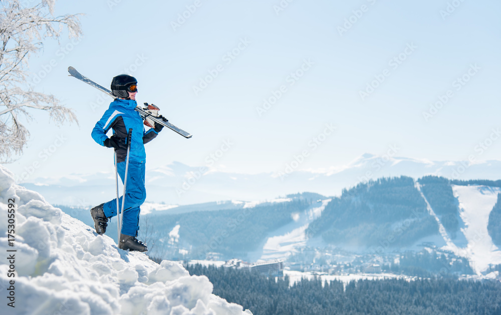 Full length shot of a female skier walking down the slope with her skis on the shoulder, copyspace winter ski resort and mountains on the background