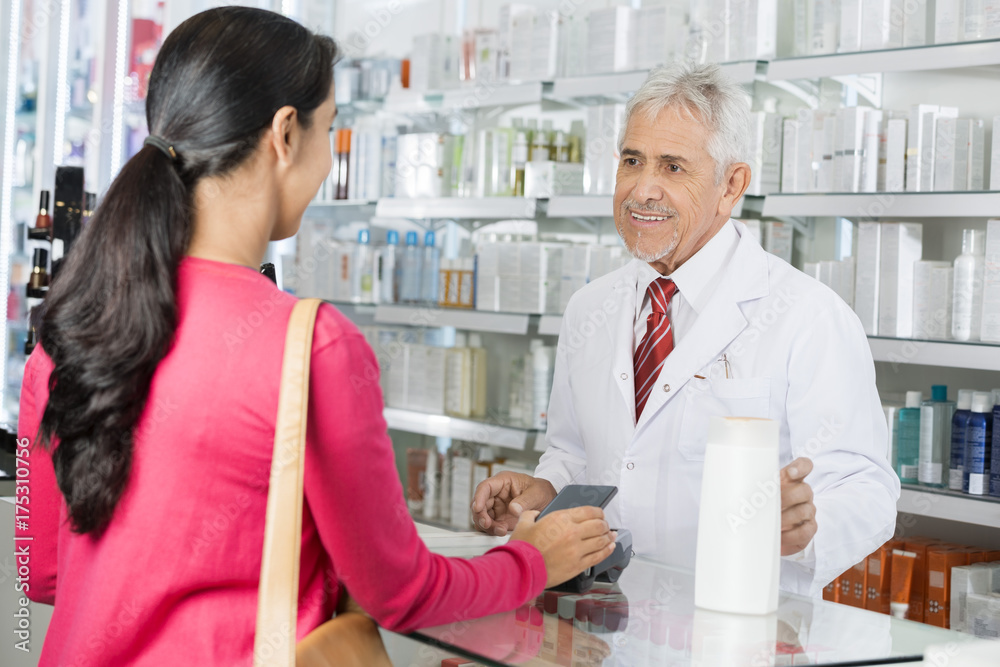 Pharmacist Looking At Female Making NFC Payment For Shampoo