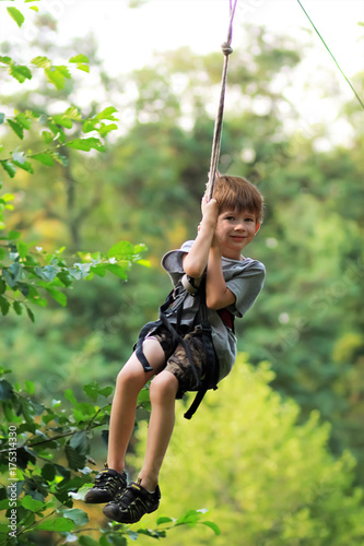 Happy boy successfully passed an obstacle course and flying in the air on zip line in grey t shirt and shorts in the summer amusement rope park among trees and lush foliage