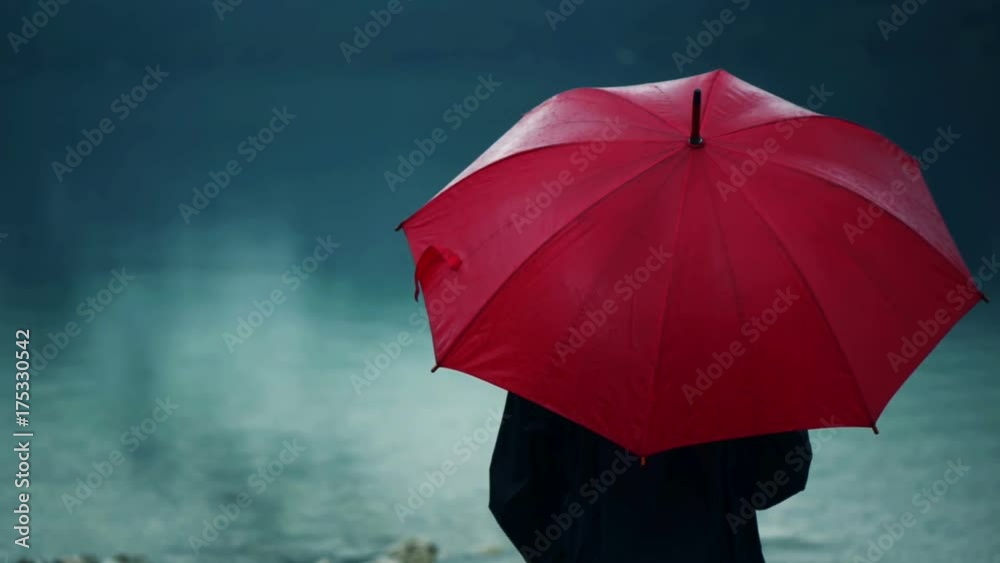 Woman with red umbrella contemplates on rain in front of a lake. Sad and lonely female person looking into distance.