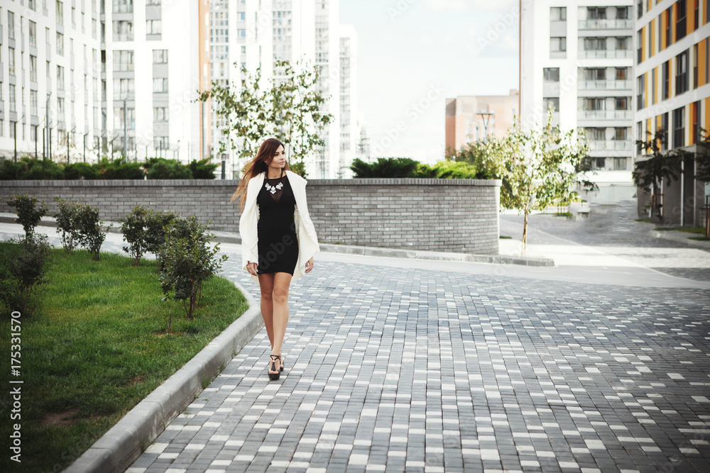 Beautiful young fashionable woman in a black short dress and white cardigan posing in the city. Fashion photo