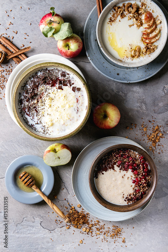 Variety bowls of milk cereal porridge with different additives, served with apples, berries and seeds over gray kitchen table. Top view with space