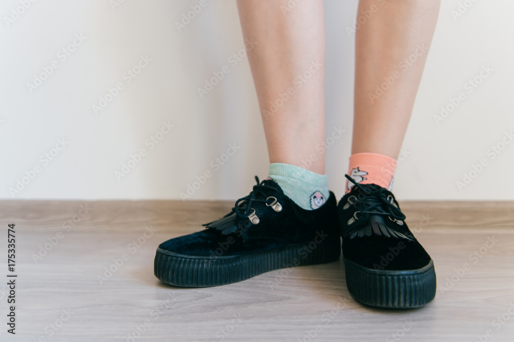 Female feet in black elegant feminine black suede shoes with tankette. Women`s legs in missmatched cotton socks. Taned soft skin. Unusual inattentive unrecognizable girl standing on floor at home.