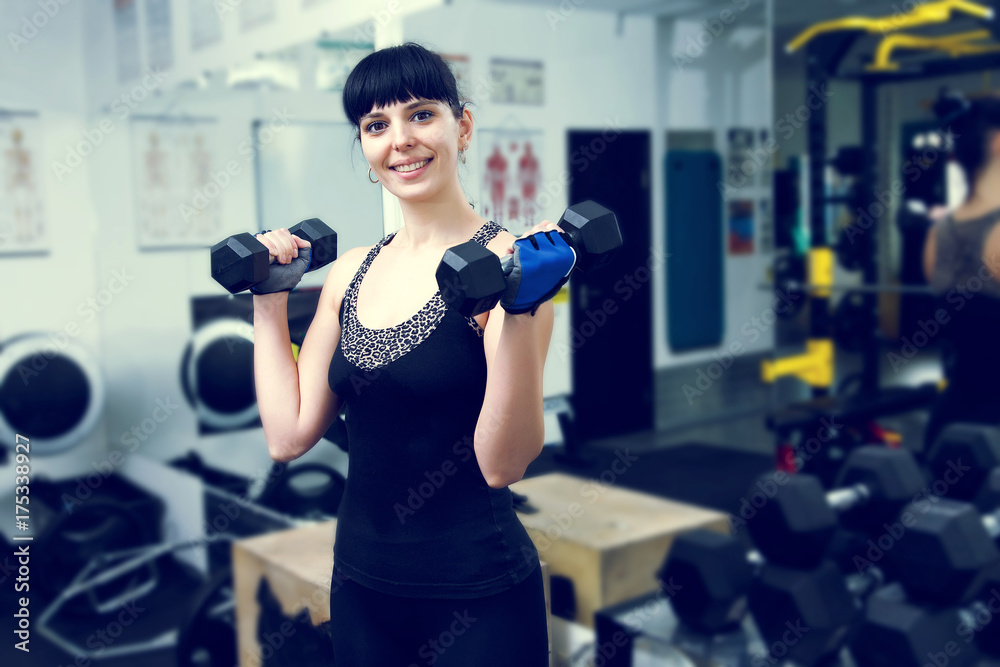 Fototapeta premium An adult woman holding dumbbell lifting up. Client fitness center. Young woman doing workout with weights on the fitness gym background. Woman working out with dumbbells in gym. Sport modern woman.