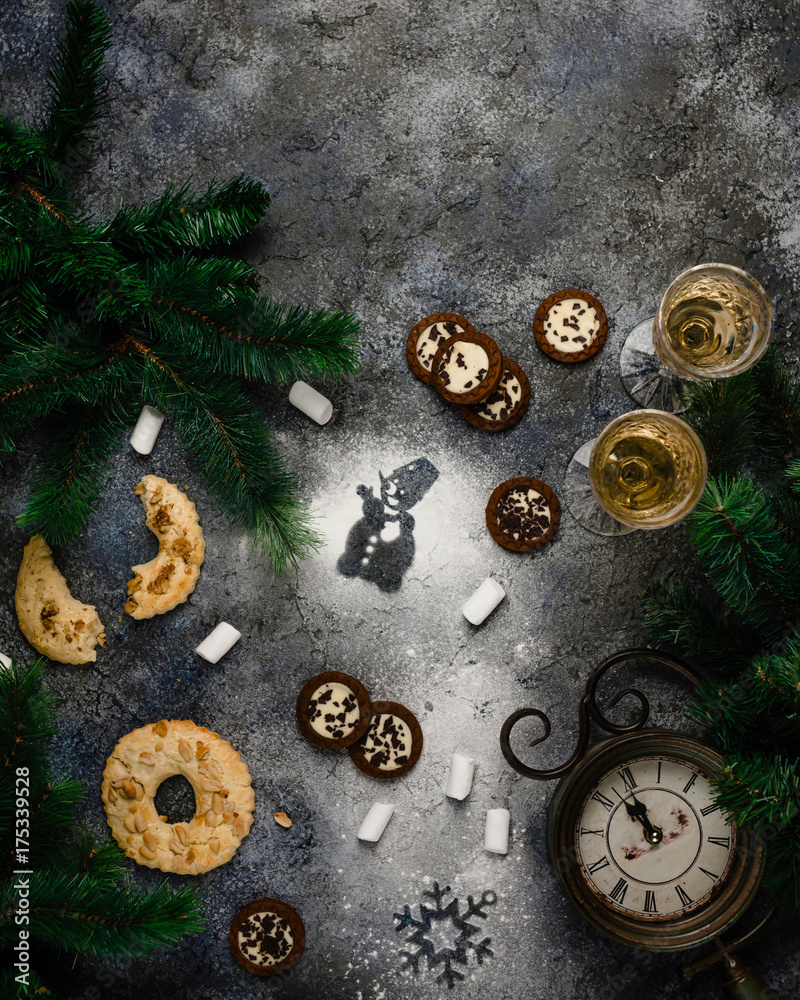 assorted cookies, two glаsses of champagne, snowman and snowflake prints made of sugar on dark blue background with old clock showing midnight among pine branches, top view