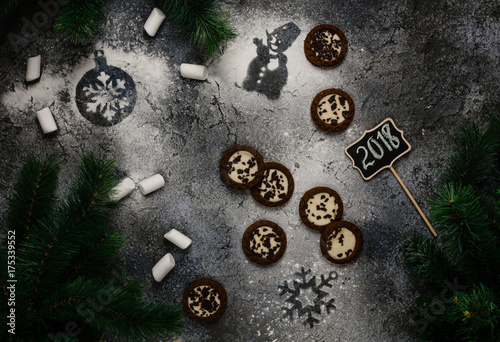 cookies and marshmallows with snowman and snowflake prints on dark blue background with 2018 writing on wooden sign among pine branches, top view