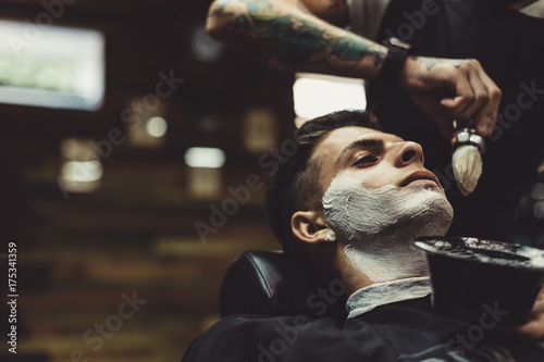 Crop stylish applying foam on customer's cheeks for shaving while working in barbershop. photo