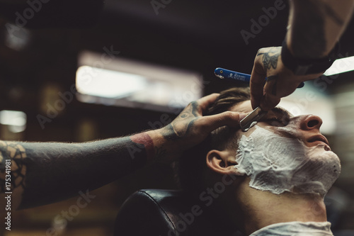 Crop stylish applying foam on customer's cheeks for shaving while working in barbershop.
