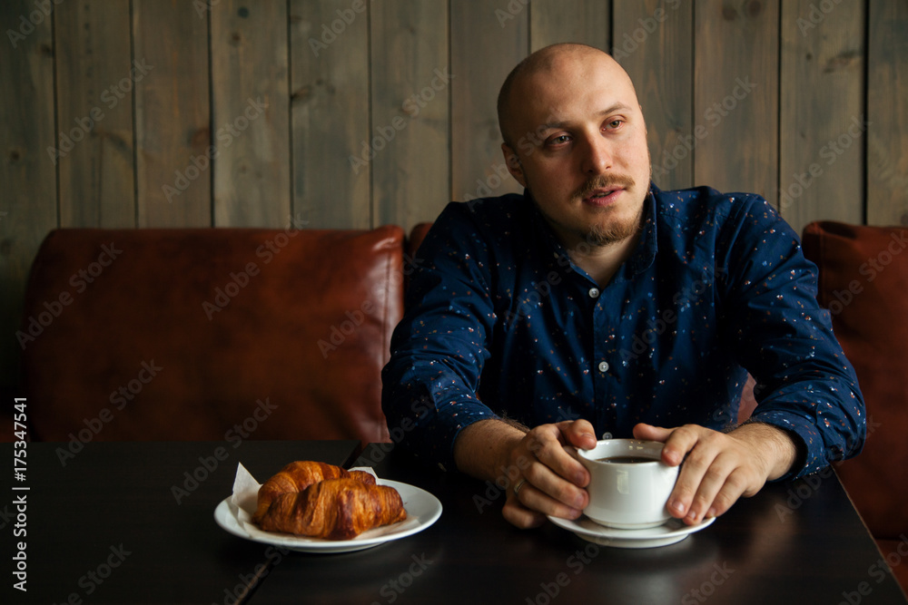 Young serious fashionable man sitting alone in loft-styled cafe and talking with somebody. Cup of coffee and croissants on the table. Former factory building, natural daylight. Young serious fashionable man sitting alone in loft-styled cafe and talking with somebody. Cup of coffee and croissants on the table. Former factory building, natural daylight.