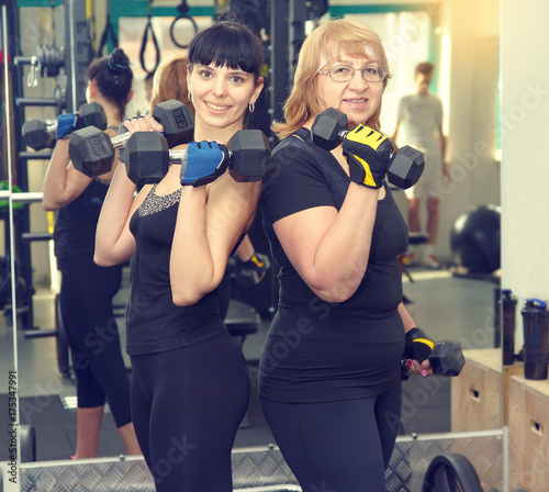 Portrait two women is engaged in a sports hall. Toned image. Women holding a dumbbell for fitness. Sporting women. Sports and fitness a lifestyle in all age. Pretty mature women. Client fitness club. © A Stock Studio