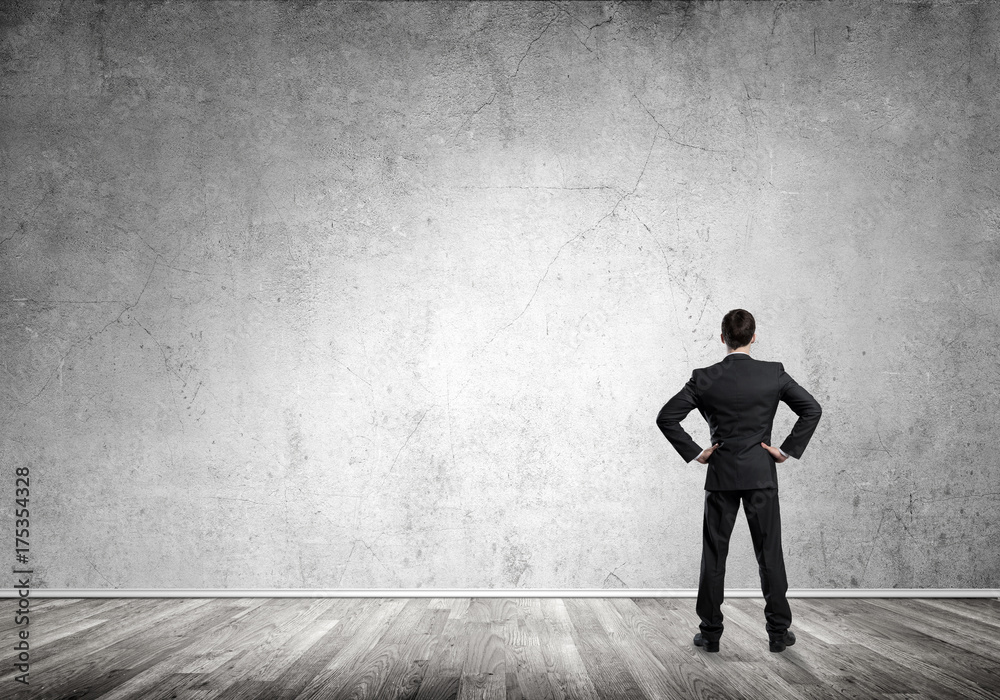 Businessman with hands on waist standing with back and looking at blank concrete wall Businessman with hands on waist standing with back and looking at blank concrete wall