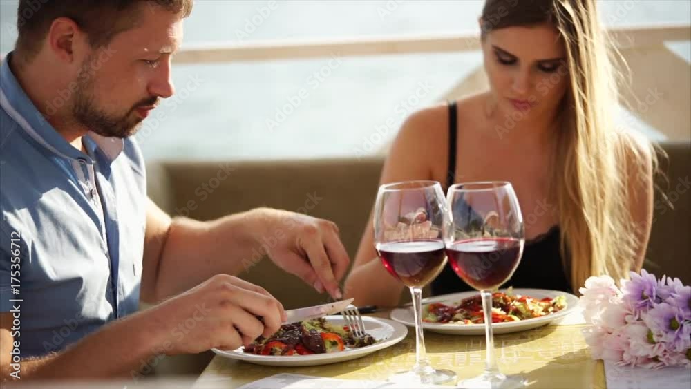 A young couple is eating dinner in a cafe during the day, a man and a woman eat tasty salads and drink wine from glasses near the river, they use forks and knives A young couple is eating dinner in a cafe during the day, a man and a woman eat tasty salads and drink wine from glasses near the river, they use forks and knives