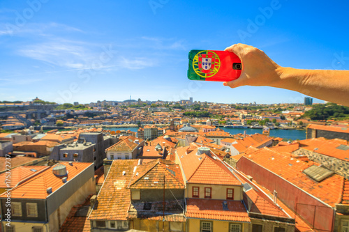 Woman takes photo of Oporto skyline from Miradouro da Vitoria by mobile phone with Portugal flag cover. Dom Luis I on Douro River on blurred background. Tourism and travel in Portugal.