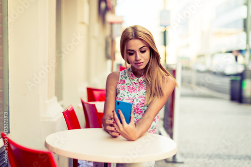 Girl blonde is sitting at an empty table of a street cafe with a smartphone in her hand