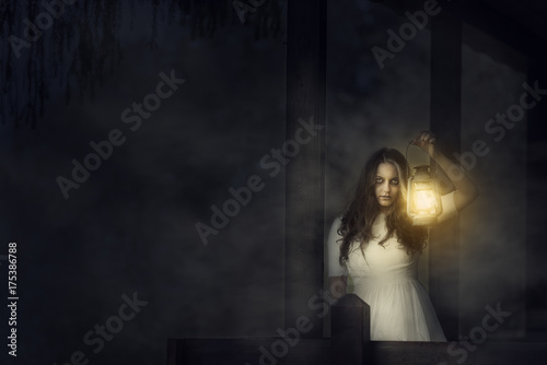 Scary woman with a lantern in night scene - Spooky image of a scary woman with dark eyes and appearance of a witch, in a white dress, holding a lit lantern, in a dark night atmosphere.