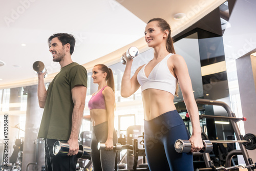 Canvas-taulu Low-angle view of three young people smiling while alternating dumbbell bicep cu