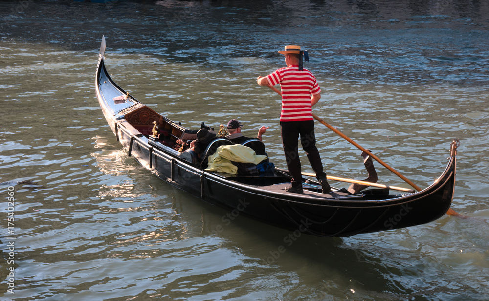 Fototapeta premium Gondola i gondolier w Wenecji / Typowy i kultowy wizerunek Wenecji, Włochy, Gondolier prowadzi swoją gondolę z dwoma turystami w piękny słoneczny dzień
