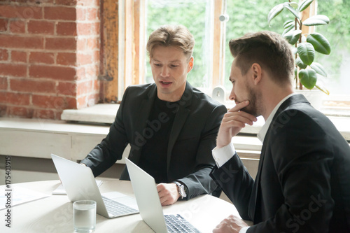 Two male executives looking at laptop screen during business meeting. Businessmen discuss corporate goals in front of laptops at workplace in office. Analyzing marketing strategies. Teamwork concept.