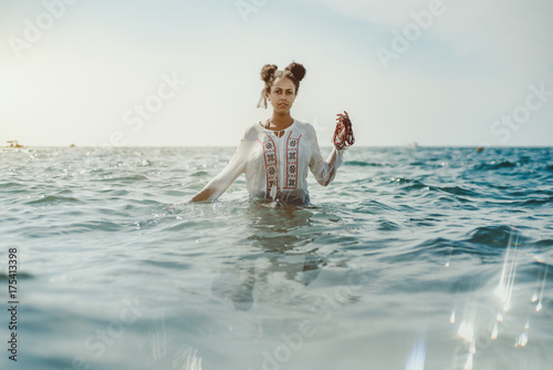 Charming young black curly lady in traditional African chemise standing up to the waist in teal seawater and holding beads in her left hand, strong reflection in the bottom, ripples on water surface photo