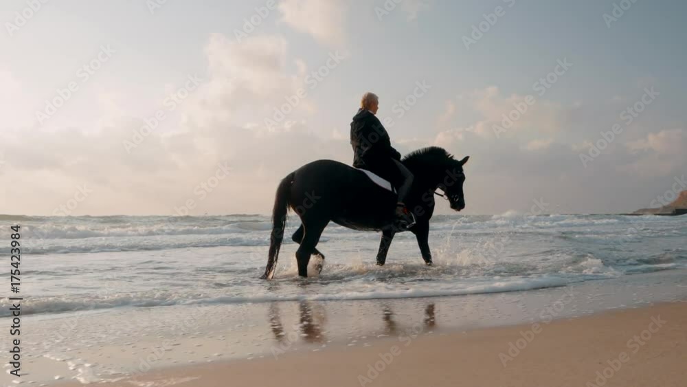 Middle aged woman with short haircut is carefully riding black horse on the seashore. Slow motion. Autumn sunrise or sunset on sea beach. Beautiful scene. Middle aged woman with short haircut is carefully riding black horse on the seashore. Slow motion. Autumn sunrise or sunset on sea beach. Beautiful scene.