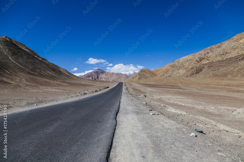 A concrete road towards beautiful rocky mountains and blue sky with peaks of Himalaya, Leh, Ladakh, Jammu and Kashmir, India