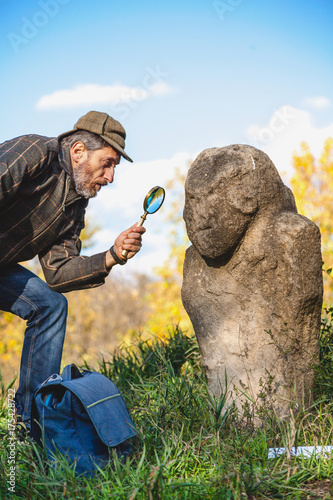 Scientific historian examines through magnifying glass stone sculpture on mound photo