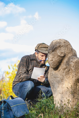 Scientific historian examines through magnifying glass stone sculpture on mound photo